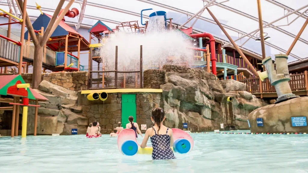 Child with float approaching splash zone under a giant tipping water bucket at Alton Towers Water Park, with play structures and slides in the background