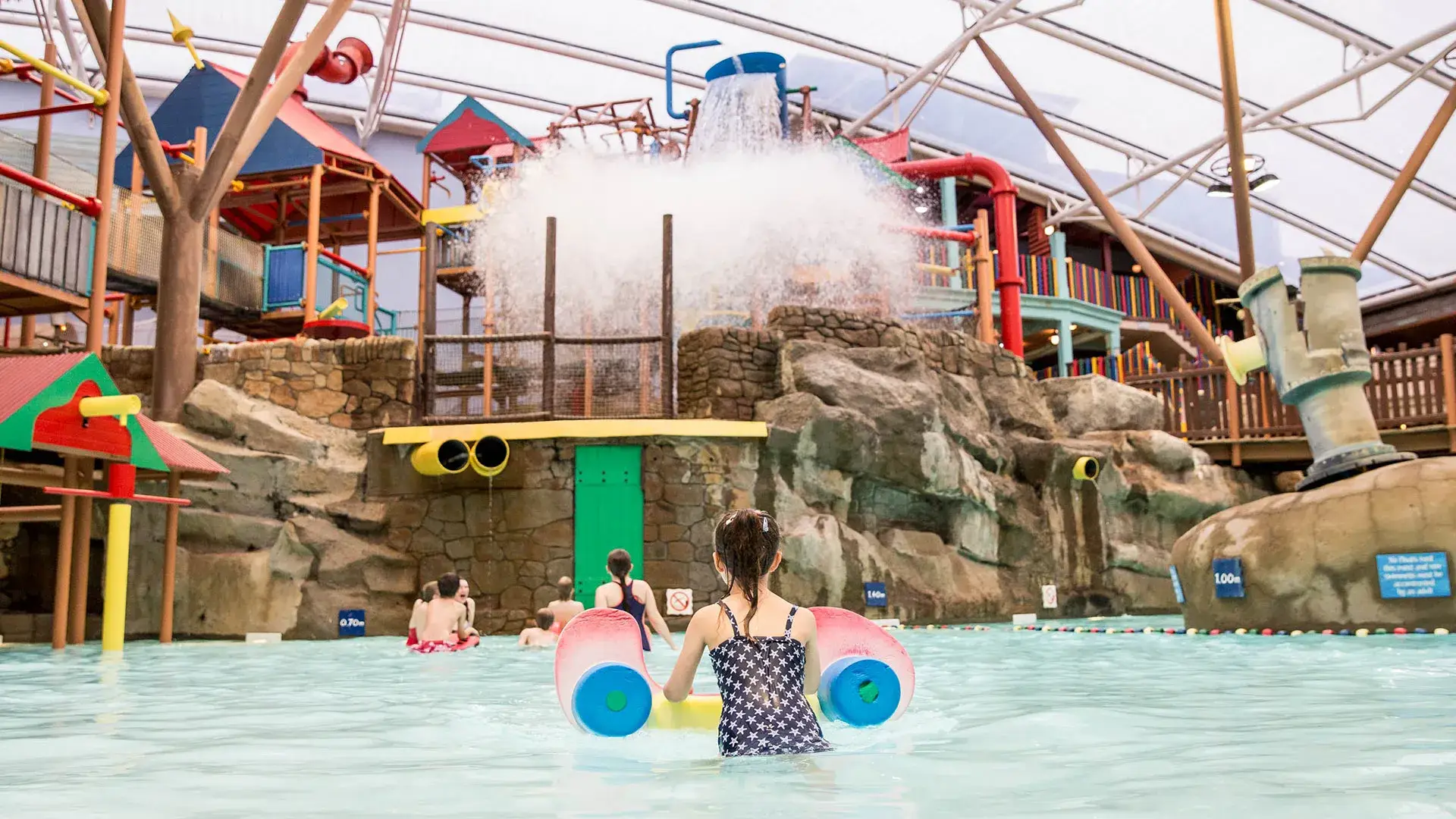 Child with float approaching splash zone under a giant tipping water bucket at Alton Towers Water Park, with play structures and slides in the background