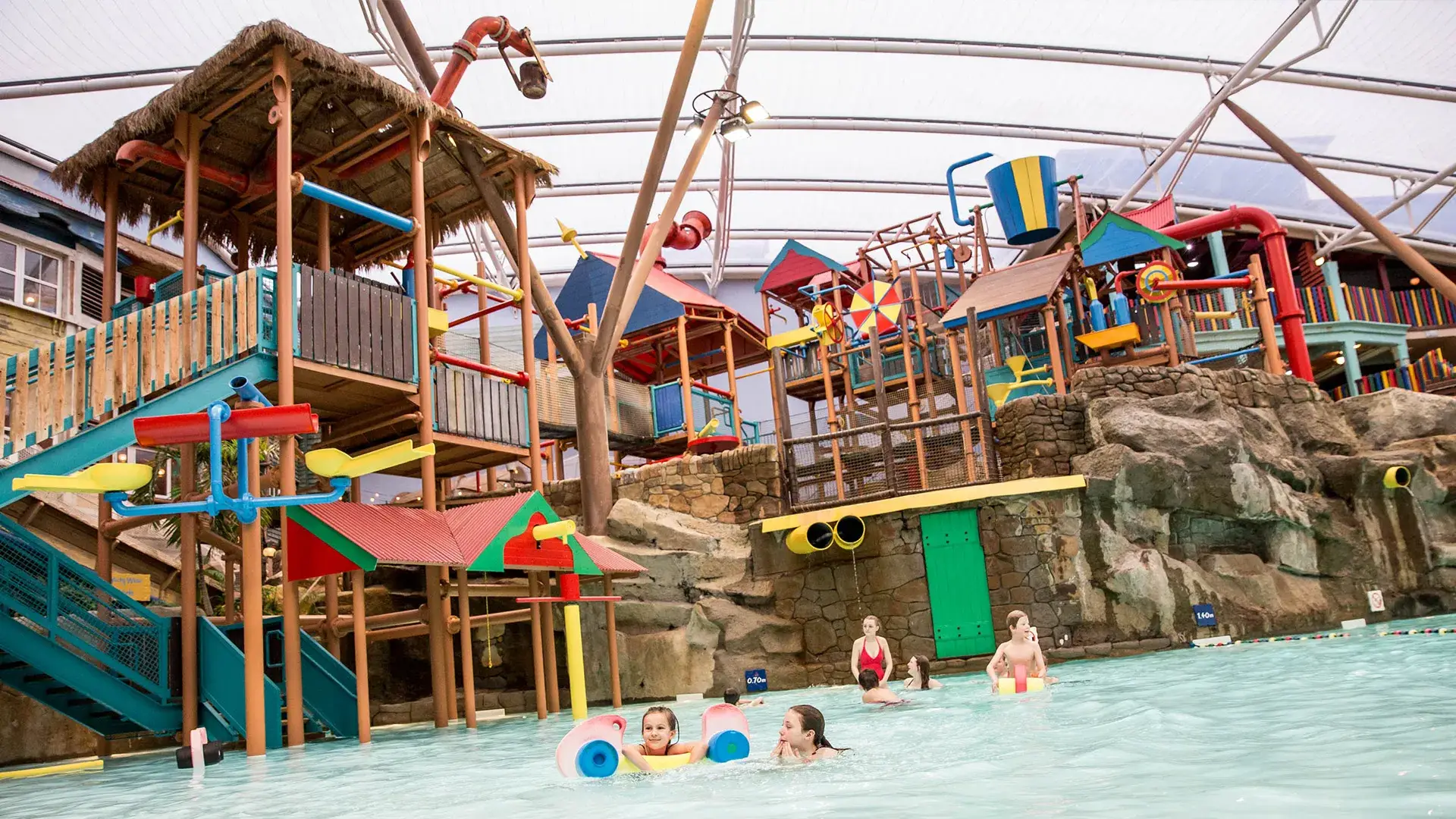 Children and families playing in the shallow pool at Alton Towers Water Park, surrounded by colourful slides, splash features, and tropical structures