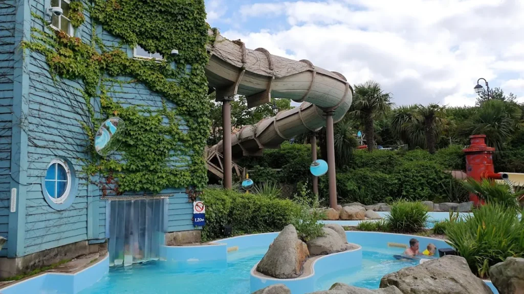 View of the outdoor section at Alton Towers Water Park in Staffordshire, showing a covered flume slide above the Calypso Creek lazy river with tropical greenery, rocks, and guests enjoying the water.