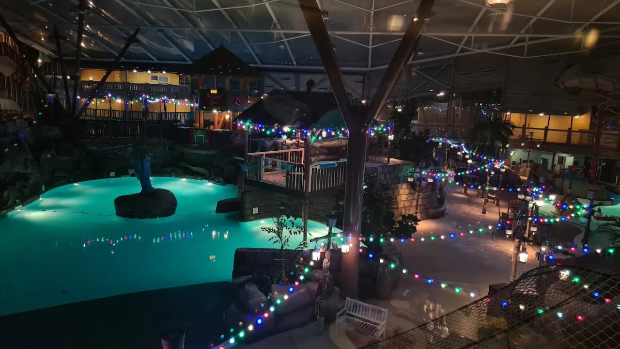 Indoor view of Alton Towers Water Park in Staffordshire, showing a leisure pool and surrounding play areas decorated with strings of colourful lights, tropical plants, and themed structures under a large glass roof.