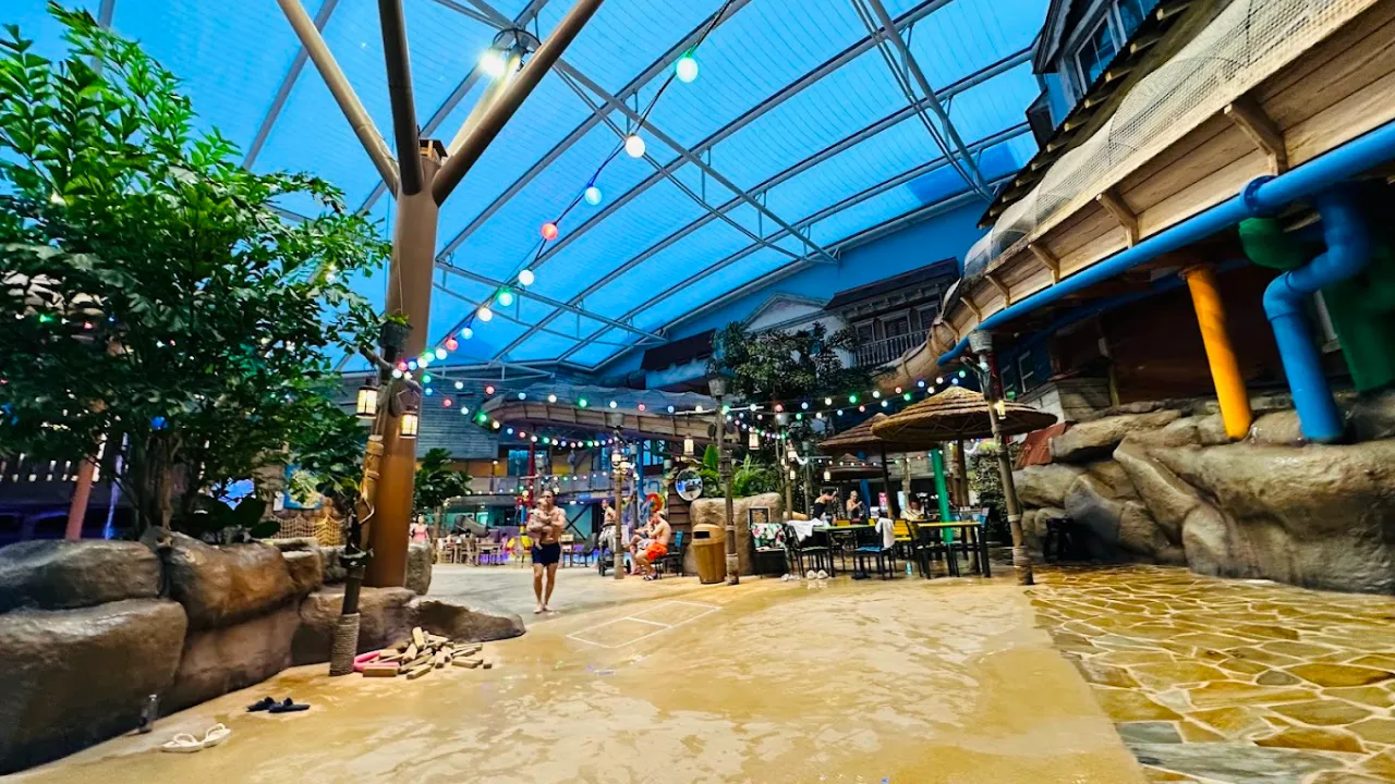 Interior view of Alton Towers Water Park in Staffordshire, showing a tropical-themed walkway with palm trees, seating areas, dining tables, and colourful string lights beneath a large glass roof.