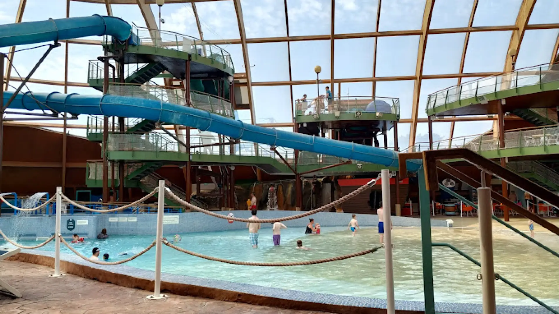 Indoor scene of Blue Lagoon Water Park showing large spiral water slides, platforms, and children playing in the shallow splash pool under a glass roof.