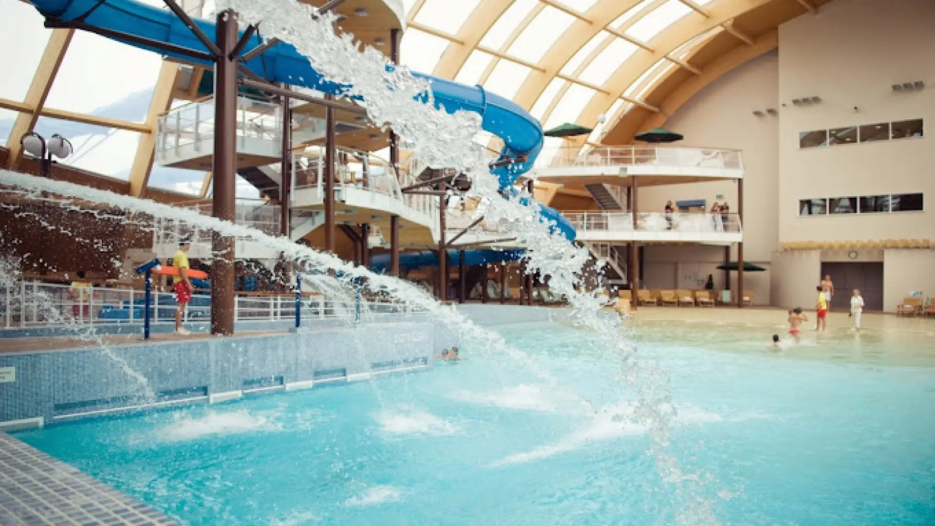 Indoor view of Blue Lagoon Water Park featuring water jets, blue slides, lifeguards, and families enjoying the shallow pool at Bluestone Resort, Wales.