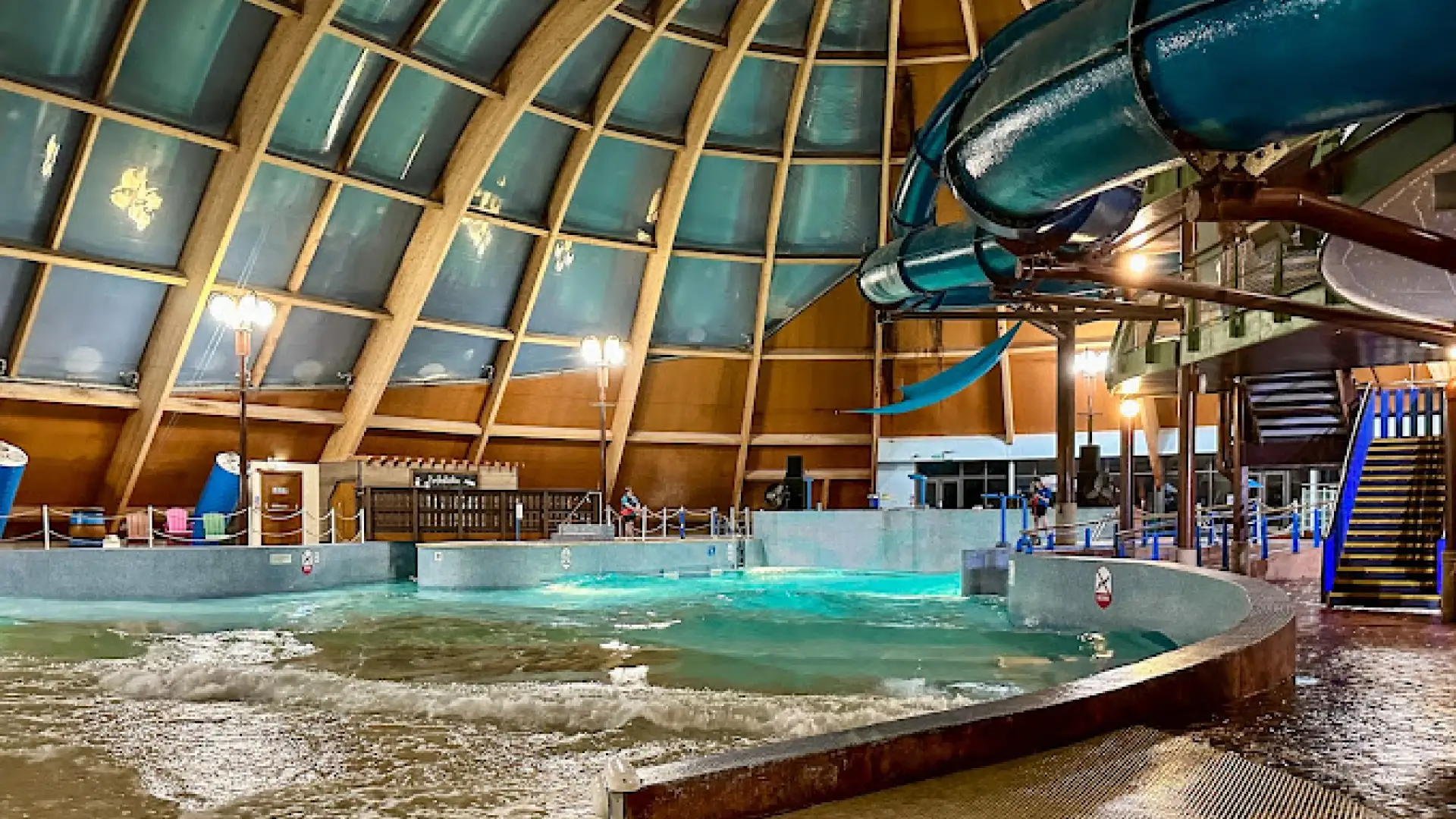 Indoor view of Blue Lagoon Water Park at night with glowing lights, wave pool in motion, and large blue water slide inside a glass-domed structure.
