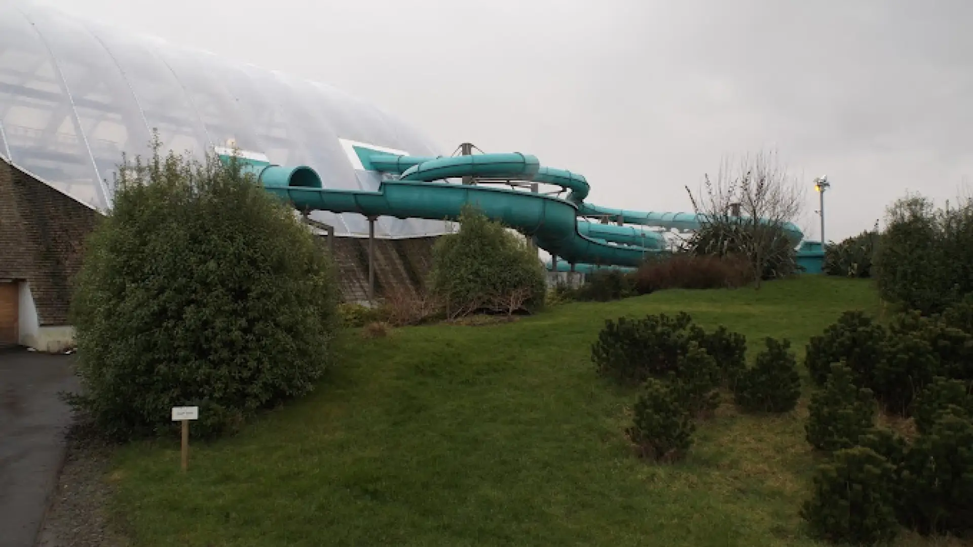 Exterior view of Blue Lagoon Water Park showing turquoise water slides emerging from the glass dome and winding through green landscaped gardens in Wales.