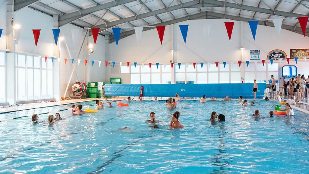 People swim and play in the spacious indoor pool at Brean Splash Waterpark, a versatile water park facility featuring a family-friendly environment with colorful floaties and poolside seating.