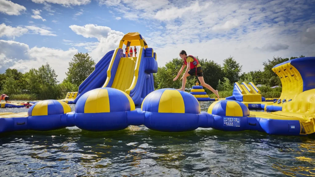 Child running across inflatable obstacle course at Cotswold Country Park and Beach on a sunny day, with bright blue and yellow water slides in the background.