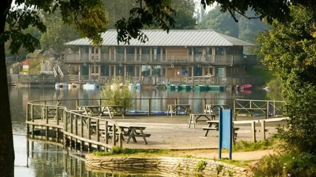 Peaceful view of wooden decking and picnic area by the lake at Cotswold Country Park and Beach, with the boat hire centre and trees in the background.