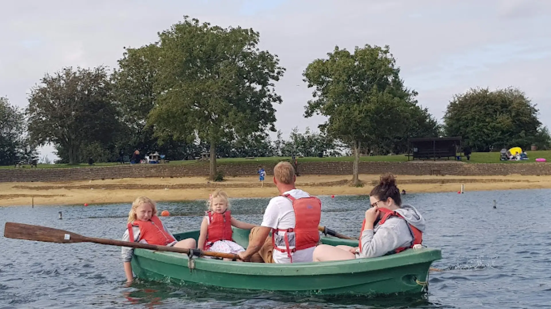 Family in a green rowboat wearing life jackets on the lake at Cotswold Country Park and Beach, with sandy shore and trees in the background.