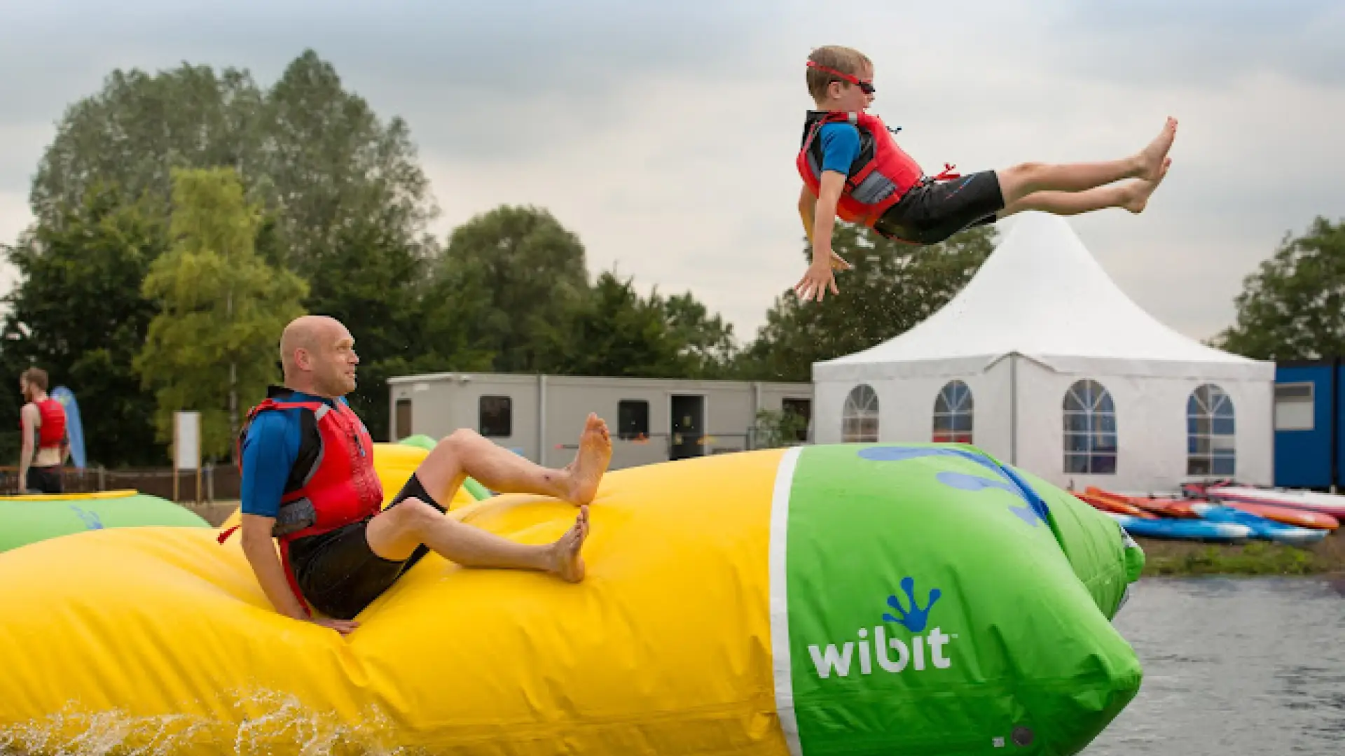 Child in red life jacket mid-air after being launched from a large inflatable blob at Cotswold Country Park and Beach, with a man seated on the blob below.