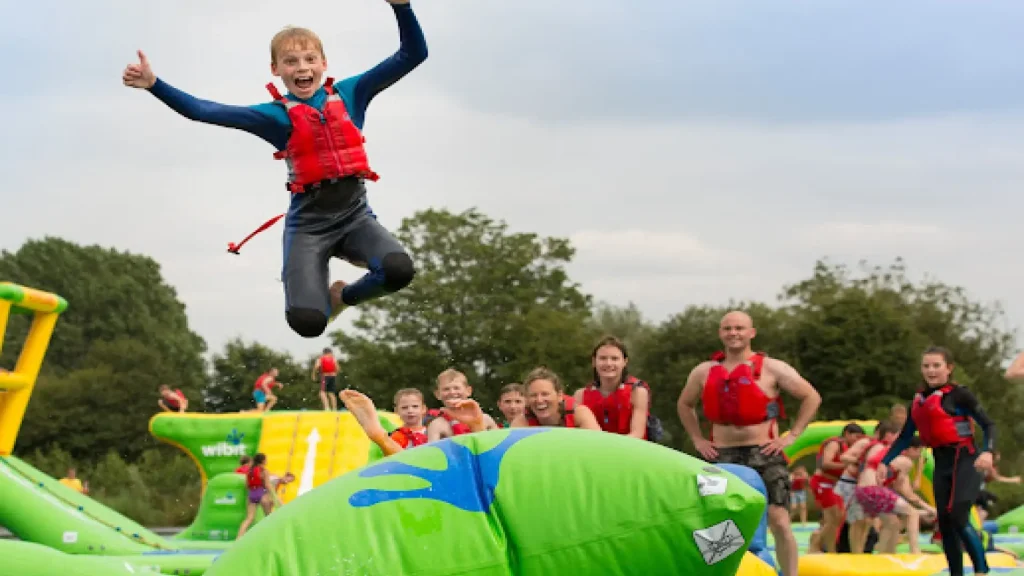 Excited child in red life jacket mid-air after jumping on an inflatable blob at Cotswold Country Park and Beach, with smiling onlookers and green inflatables behind.