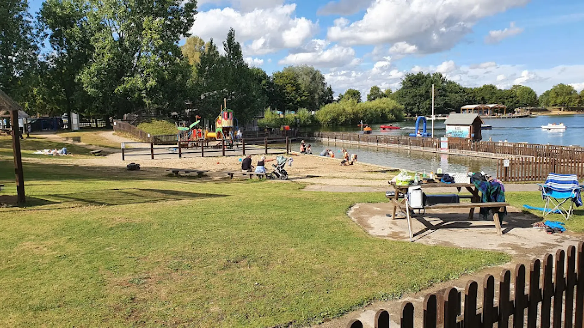 Fenced toddler play area and sandy beach beside the lake at Cotswold Country Park and Beach, with picnic benches, families, and boats in the background.