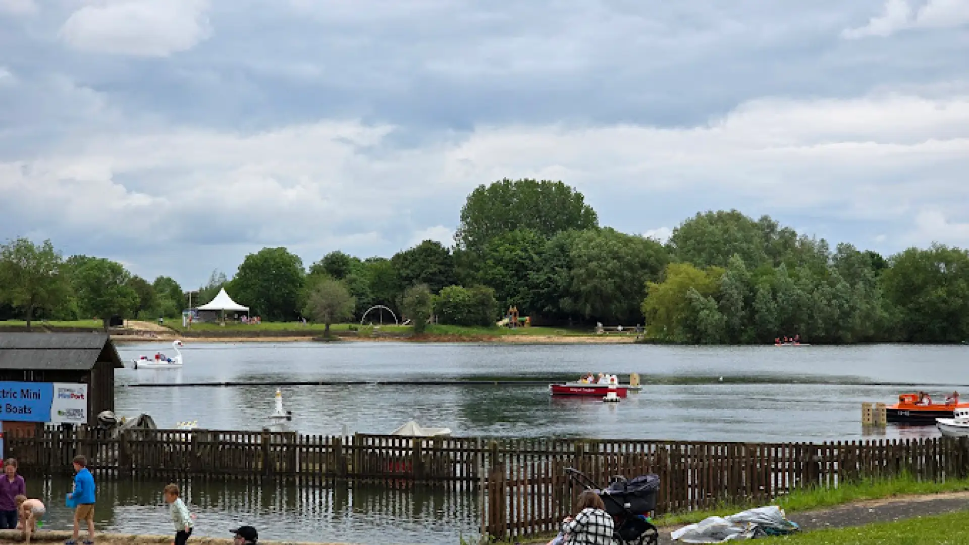 View across the lake at Cotswold Country Park and Beach showing swan pedal boats, trees, children’s play area, and families enjoying the waterfront.