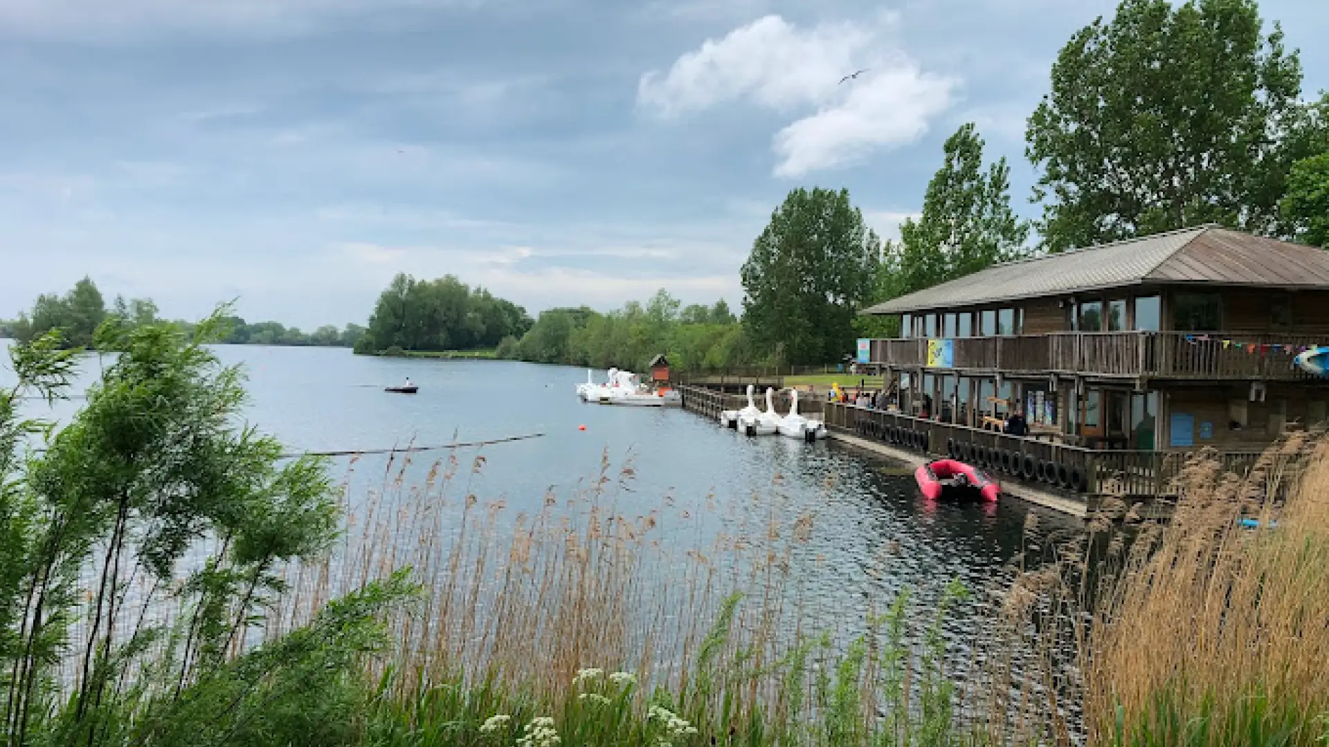 Wooden lakeside building and dock at Cotswold Country Park and Beach with swan pedal boats, paddleboats, and lush greenery surrounding the calm lake.