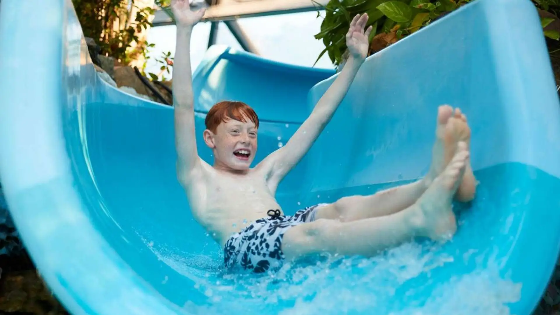 Excited child sliding down a blue water flume at the Subtropical Swimming Paradise at Center Parcs in Longleat water park, surrounded by tropical plants and splashing water.