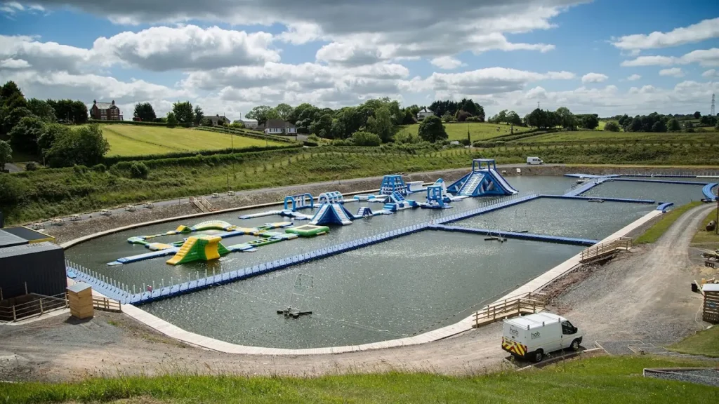 View of Let’s Go Hydro water park in Northern Ireland with blue and green inflatable obstacles, wakeboarding area, and lush green countryside in the background.