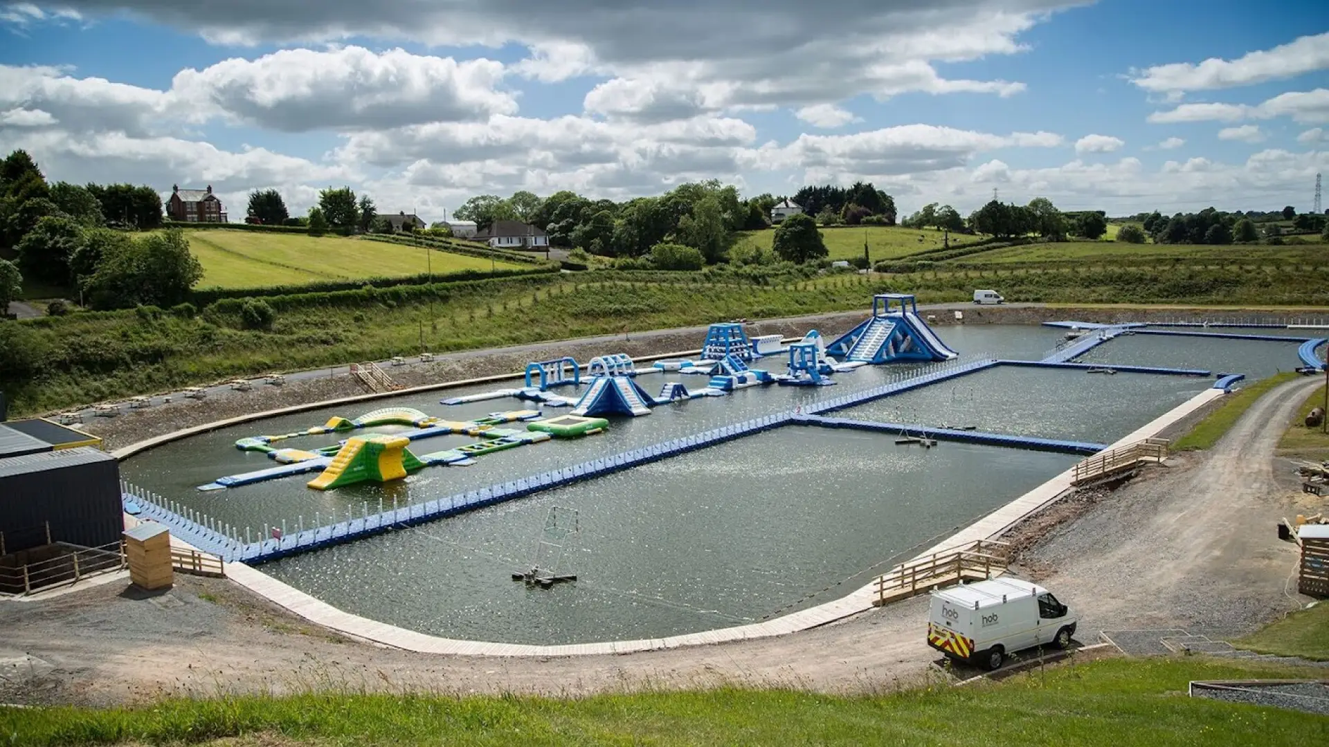 View of Let’s Go Hydro water park in Northern Ireland with blue and green inflatable obstacles, wakeboarding area, and lush green countryside in the background.