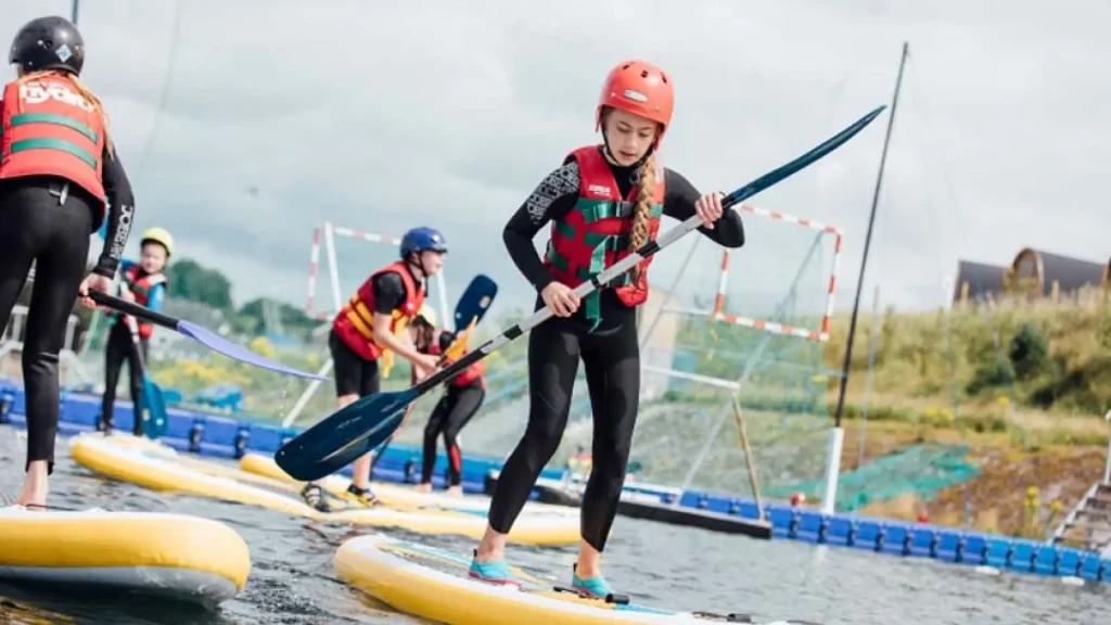Children paddleboarding in wetsuits and life jackets at Let's Go Hydro water park in Northern Ireland, with inflatable sports zones in the background.