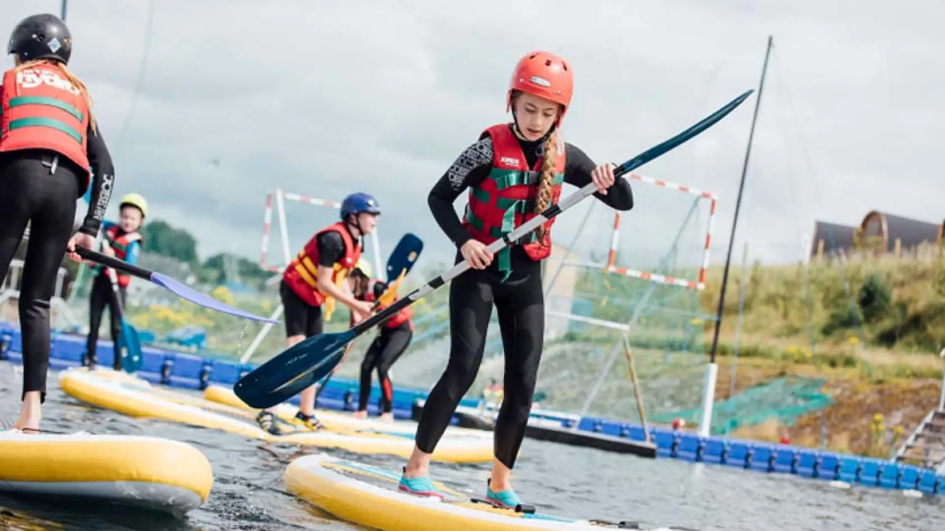 Children paddleboarding in wetsuits and life jackets at Let's Go Hydro water park in Northern Ireland, with inflatable sports zones in the background.