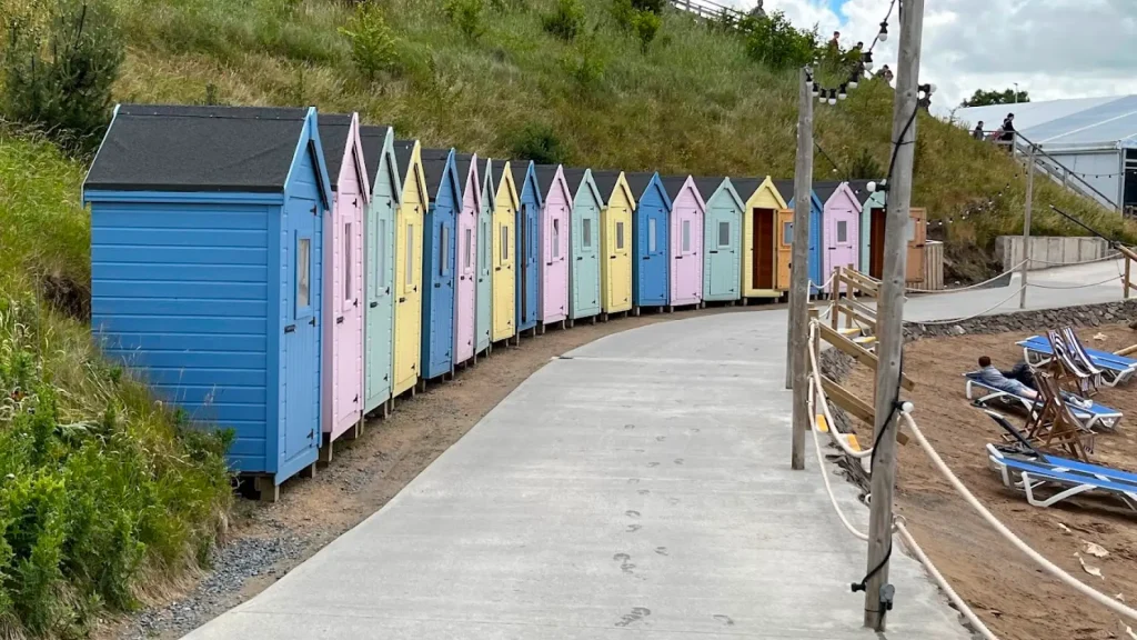 Colourful pastel beach huts lining a path beside a sandy lounge area at Let's Go Hydro water park in Northern Ireland, with deck chairs and greenery.