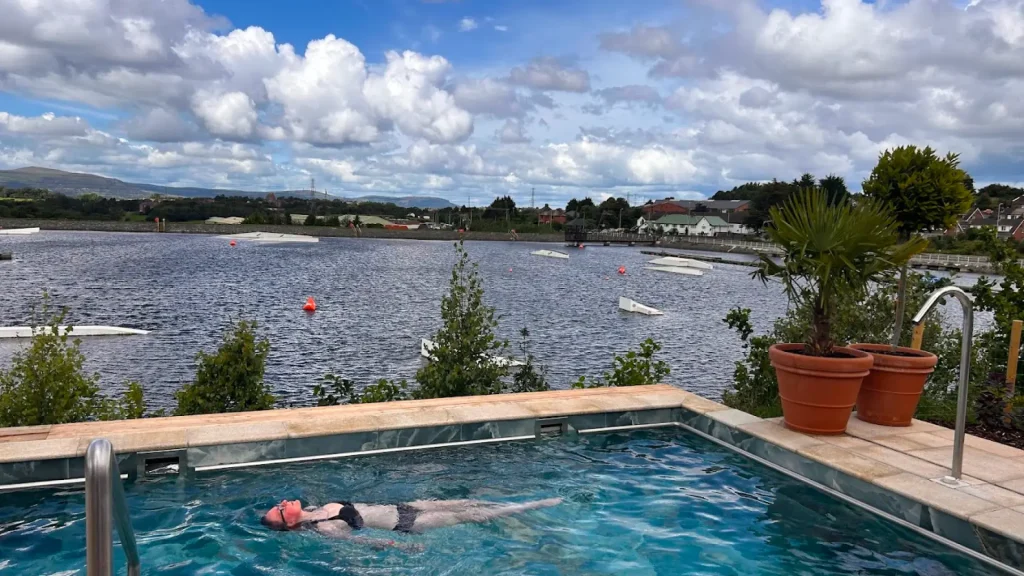 Woman relaxing in an outdoor hot tub at Let's Go Hydro, overlooking the lake and wakeboarding ramps, with palm trees and scenic views in the background.