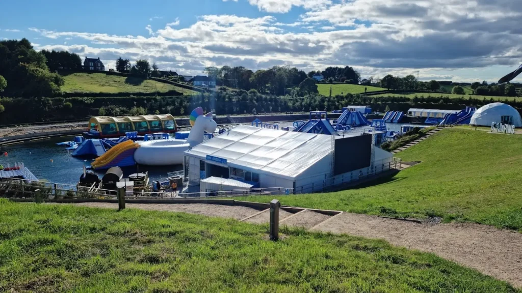 Wide shot of Let's Go Hydro featuring inflatable water attractions, a large unicorn slide, glamping dome, and outdoor activity centre surrounded by green hills.