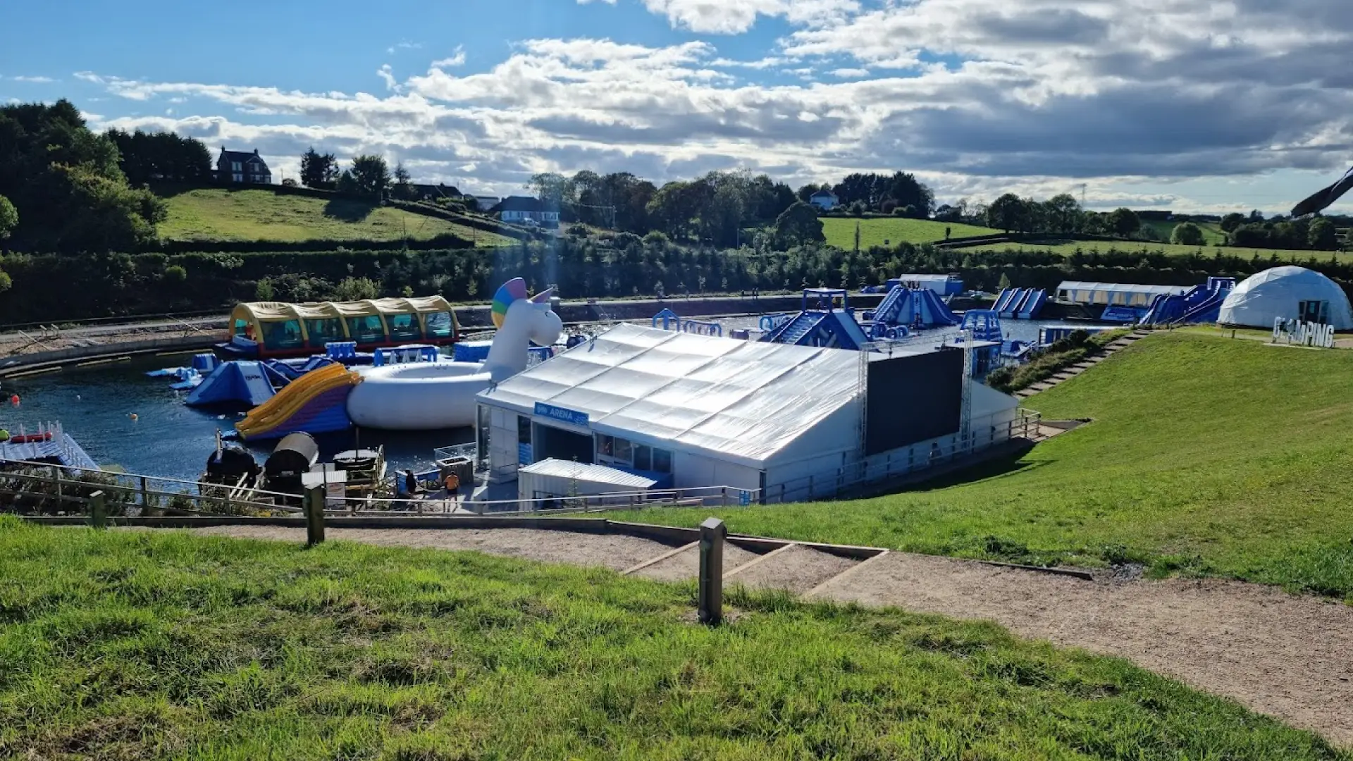 Wide shot of Let's Go Hydro featuring inflatable water attractions, a large unicorn slide, glamping dome, and outdoor activity centre surrounded by green hills.