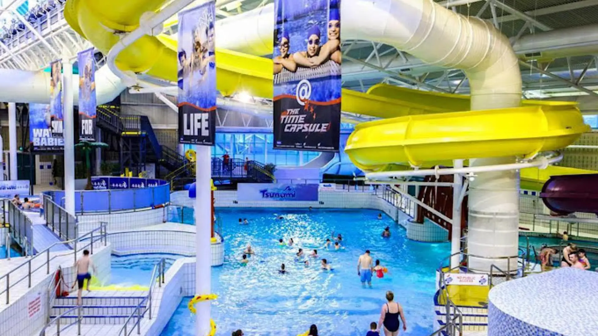Families enjoy Tsunami wave pool while overhead yellow flume twists inside The Time Capsule Water Park Scotland, Coatbridge. ​​