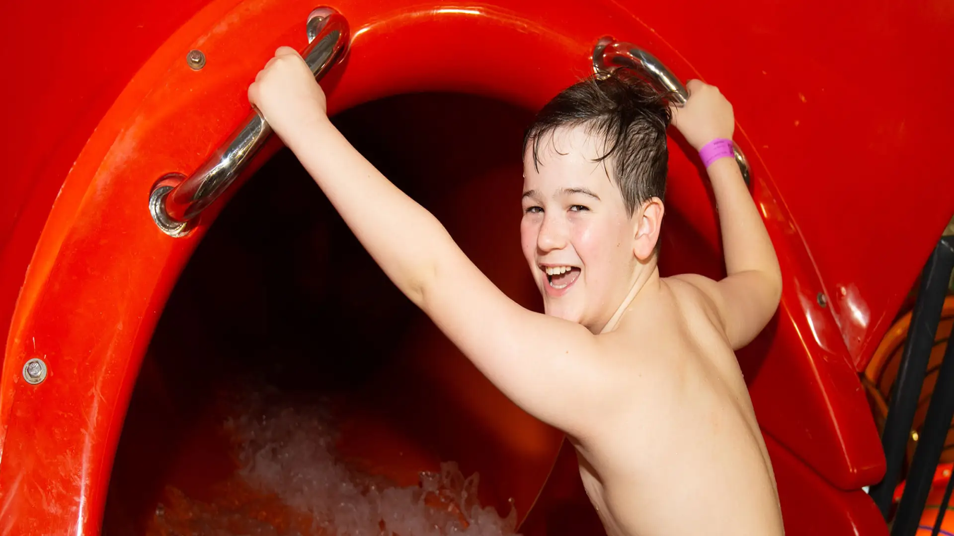 Smiling boy holding onto handles at the entrance of a red water slide at The Wave Waterpark, Coventry.
