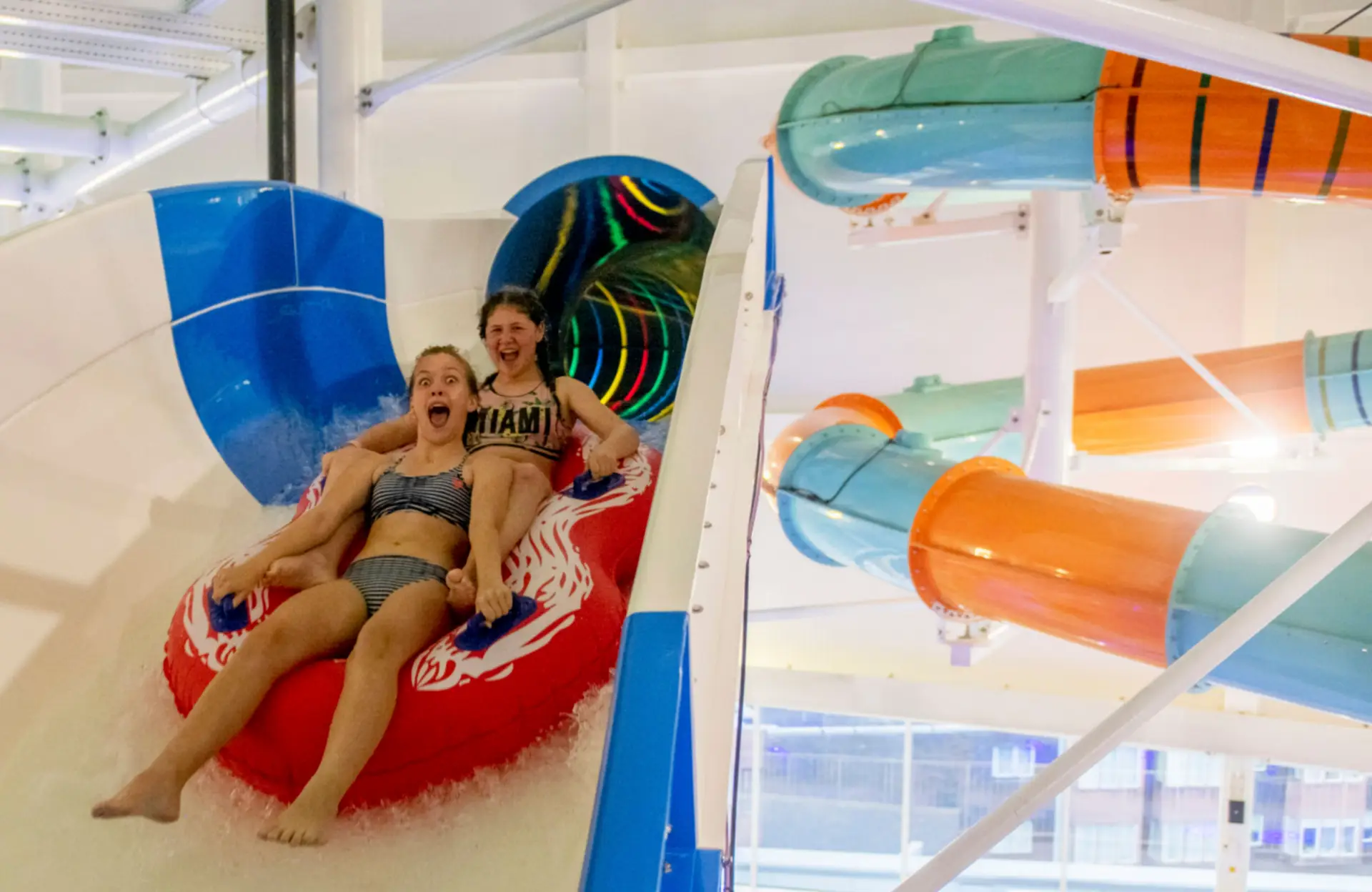 Two girls riding a red inflatable tube down a blue and white waterslide at The Wave Waterpark, with colourful slides visible in the background.
