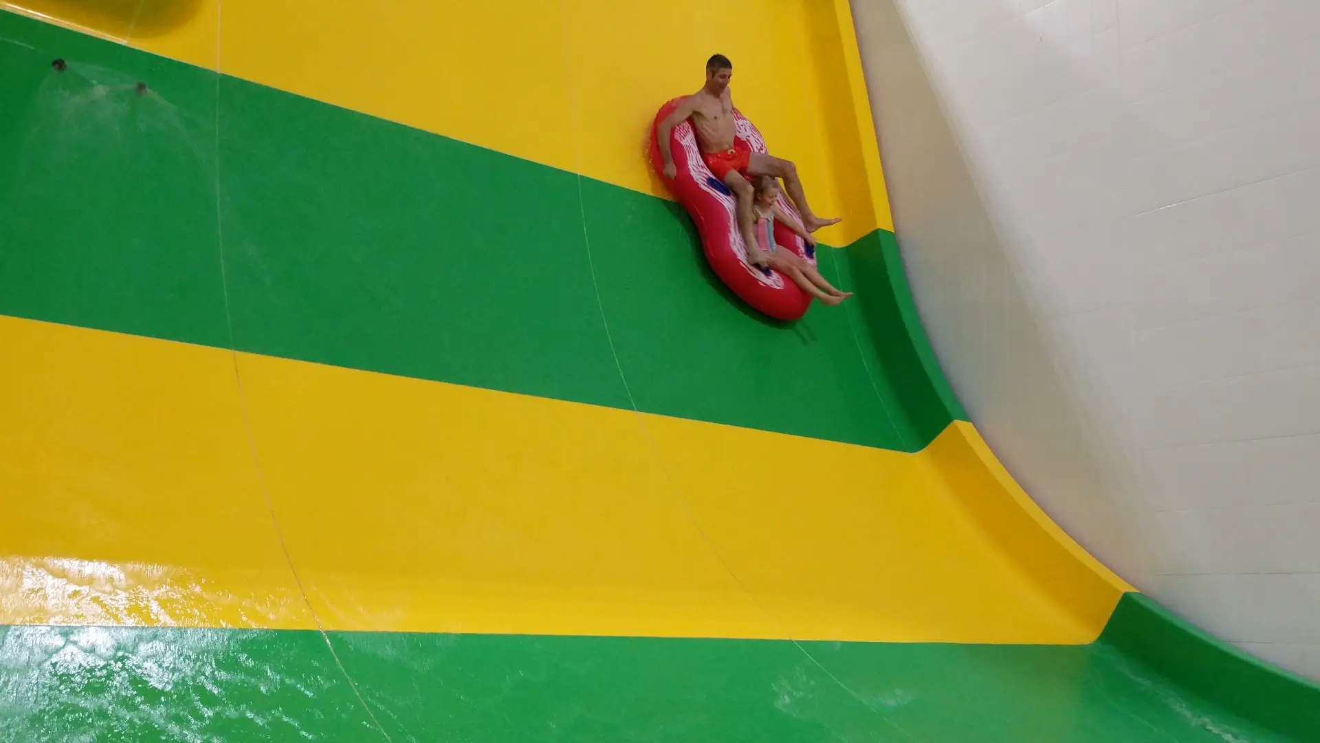Man and child on a red inflatable descending a steep green and yellow water slide at The Wave Waterpark, New Union Street, Coventry.