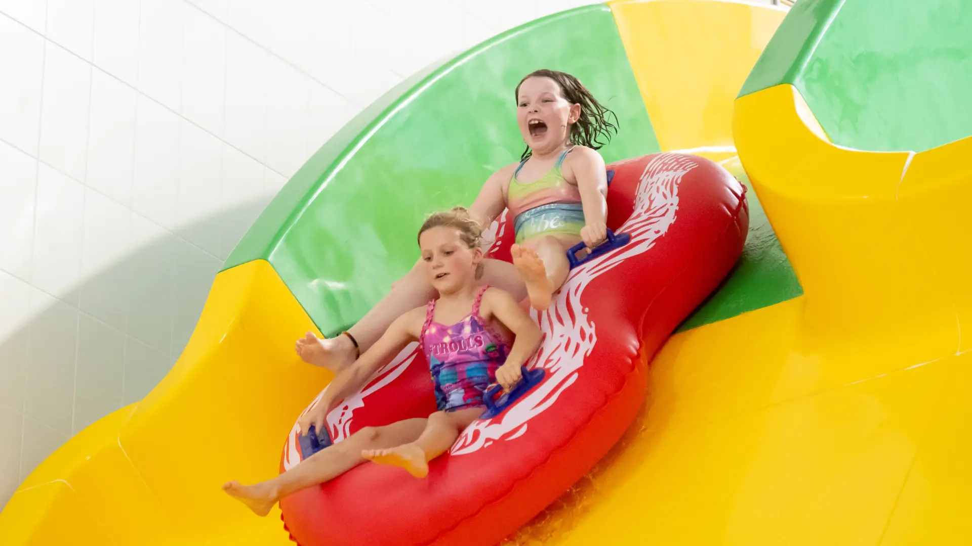 Two young girls on a red inflatable riding a yellow and green water slide at The Wave Waterpark, Coventry.
