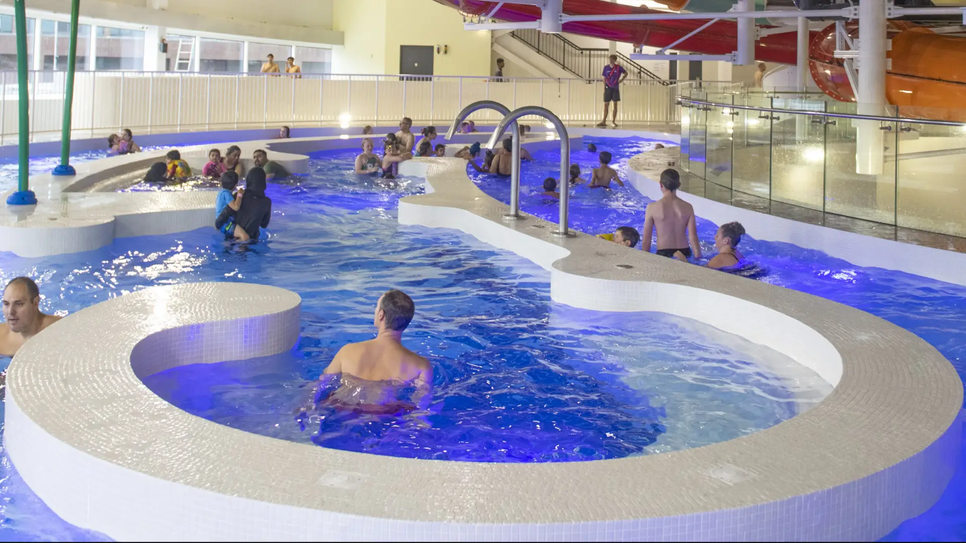 Families swimming and relaxing in a warm, blue-lit pool area at The Wave Waterpark, with waterslides and lifeguards visible in the background.
