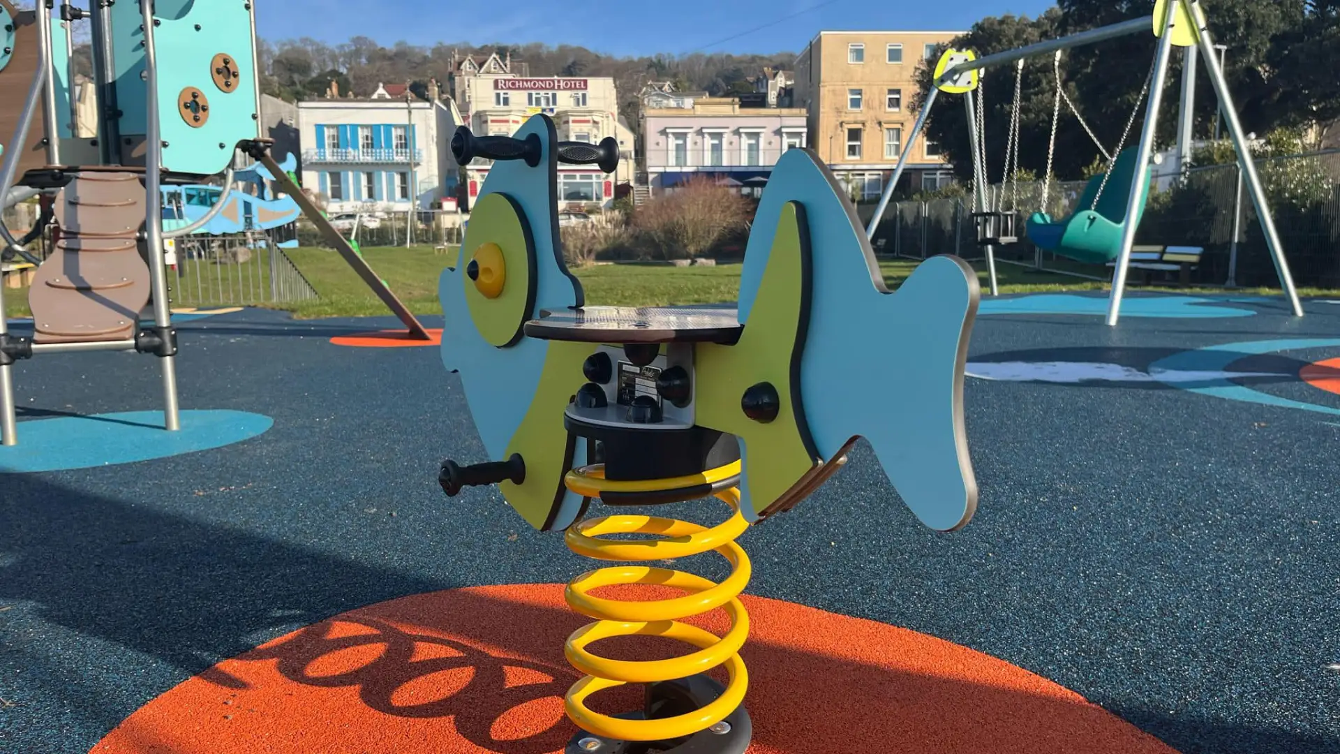 Bright fish-shaped spring rocker at Water Adventure Play Park in Weston-super-Mare Play Park, with swings and climbing frames in background.