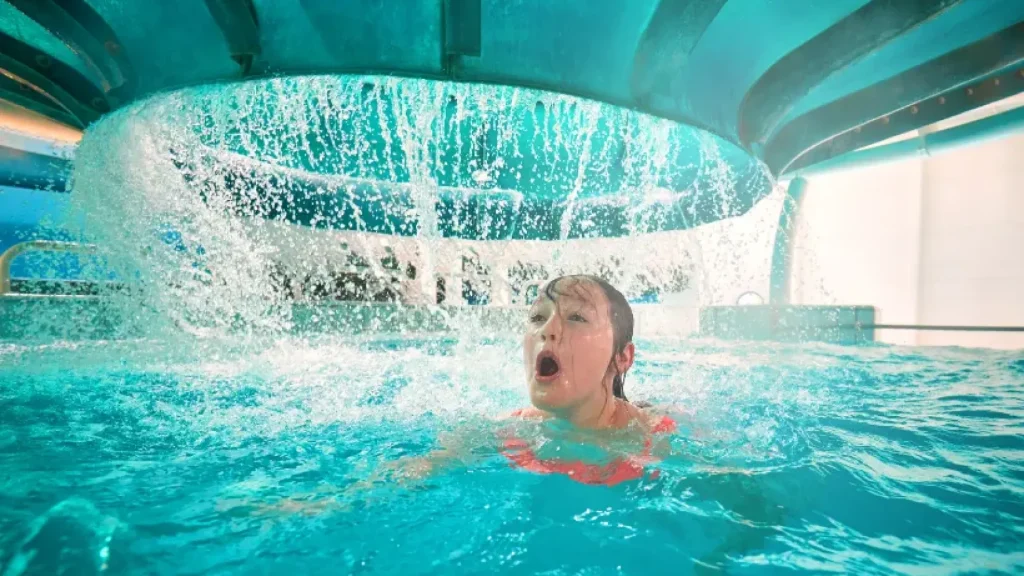 Child swimming beneath a powerful indoor water feature at Butlins Splash Waterworld Minehead water park, surrounded by turquoise water and vibrant lighting.