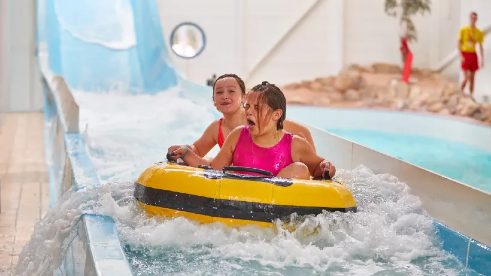 Two children riding a yellow inflatable down a water slide at Butlins Splash Waterworld Minehead water park, with visible excitement and splashing water.