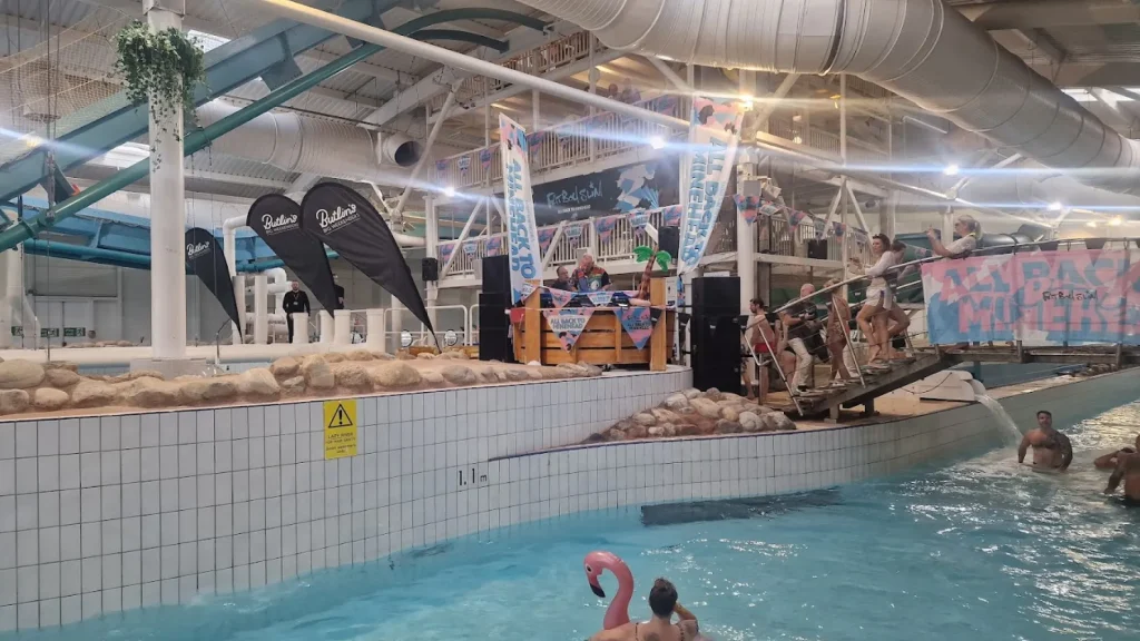 Indoor pool scene at Butlins Splash Waterworld Minehead water park featuring a live DJ setup, event flags, and guests enjoying music and swimming.