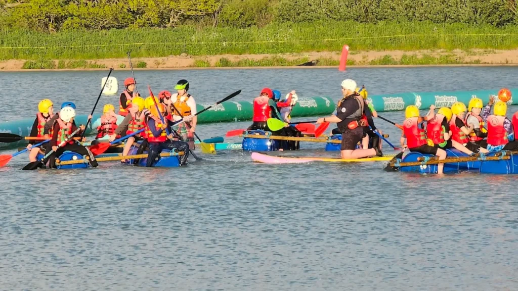Children and instructors paddling on homemade rafts during a group activity session at West Country Water Park, wearing life jackets and helmets.