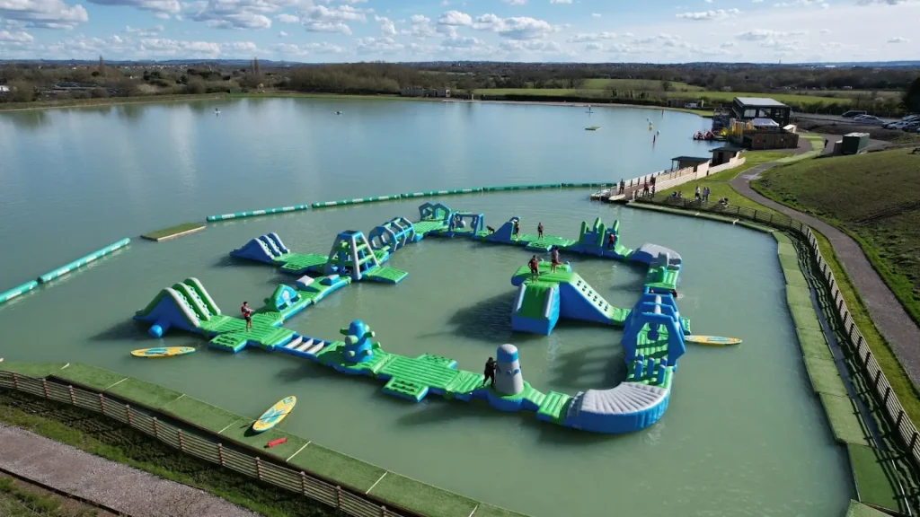 Aerial view of the inflatable aqua park course at West Country Water Park, featuring climbing walls, slides, and paddleboards on a large freshwater lake.