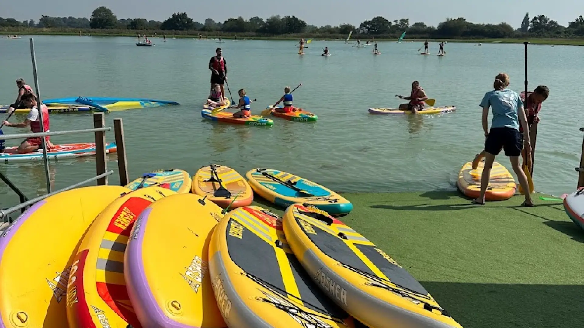 Families and children paddleboarding on a sunny day at West Country Water Park, with colorful paddleboards stacked by the lakeside launch area.