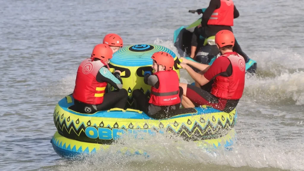 Group of people riding an inflatable water tube at high speed, wearing helmets and life vests during a water sports activity at West Country Water Park.