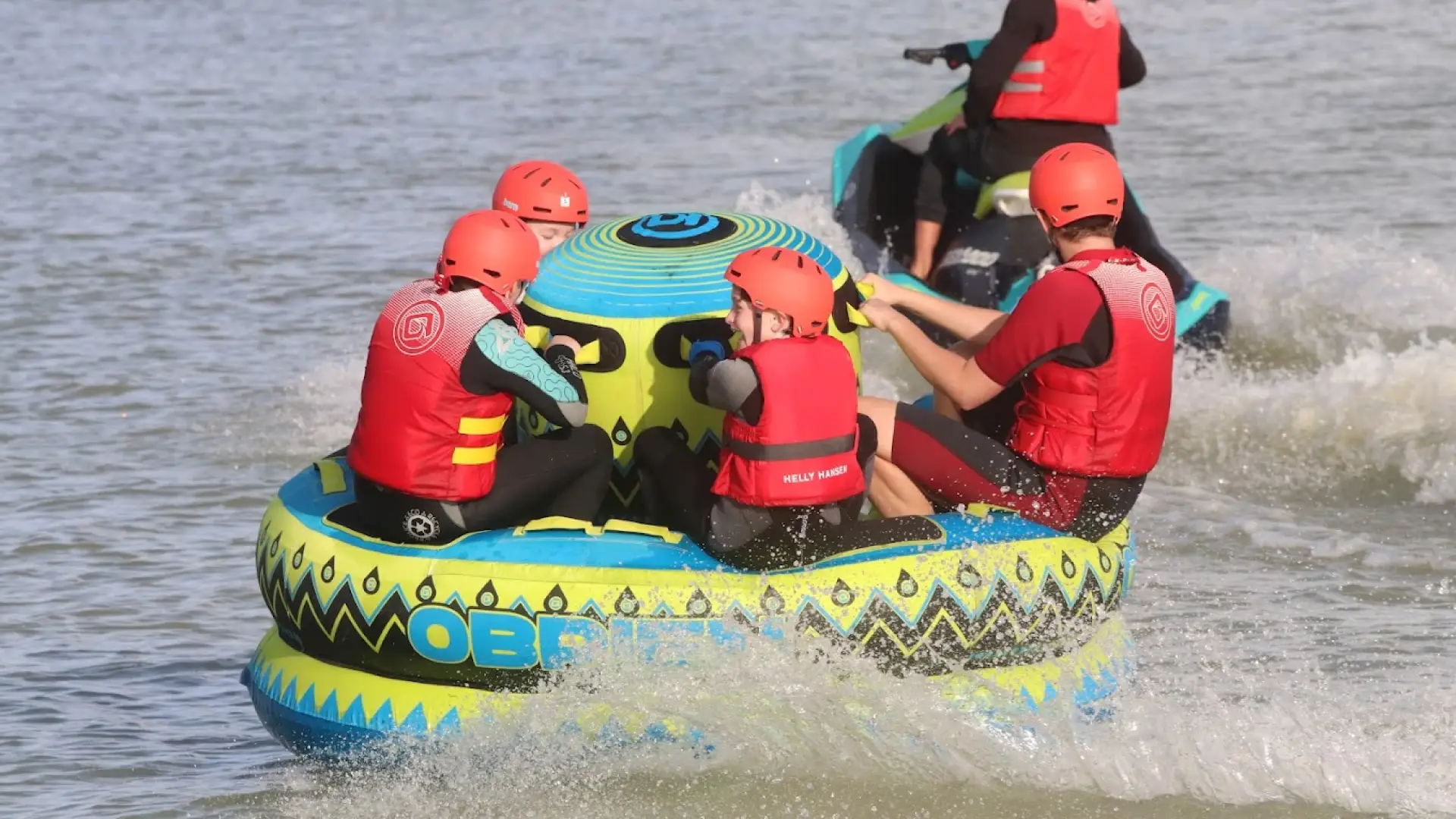 Group of people riding an inflatable water tube at high speed, wearing helmets and life vests during a water sports activity at West Country Water Park.