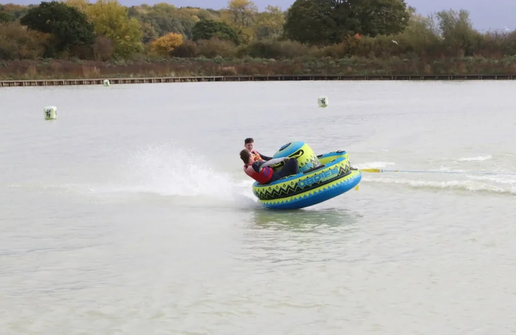 Two people riding an inflatable towable tube at high speed across the lake at West Country Water Park, creating waves and splashes in motion.