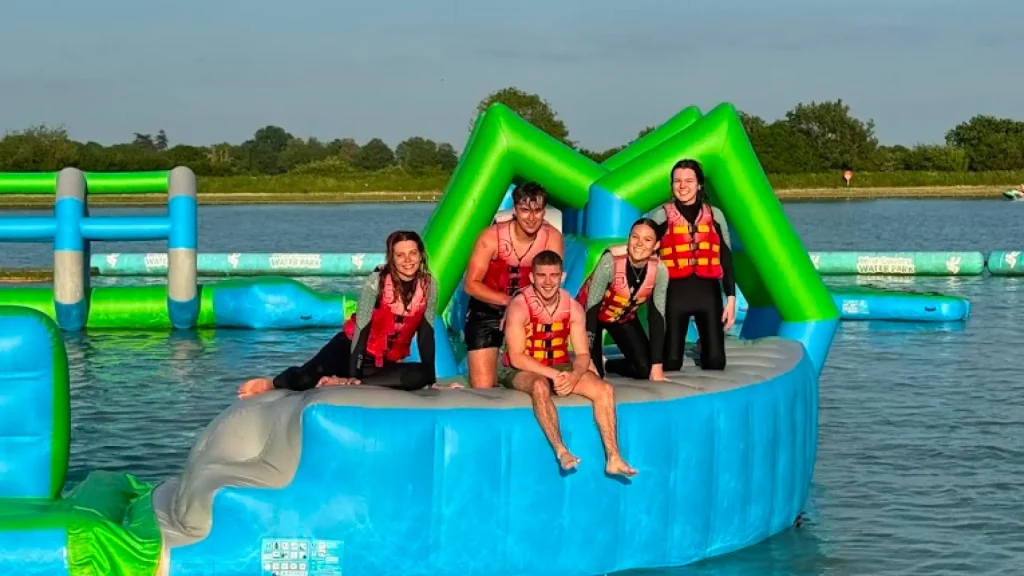 Group of young adults in life vests posing on a floating obstacle at the inflatable aqua park at West Country Water Park on a sunny day.