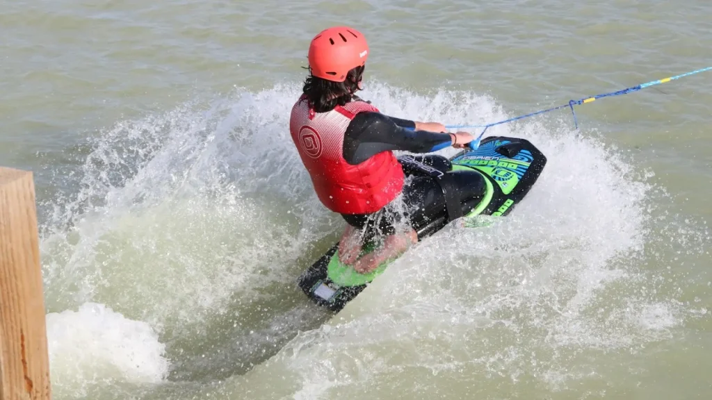 Wakeboarder carving through the water at high speed while being towed, wearing a helmet and life vest at West Country Water Park.