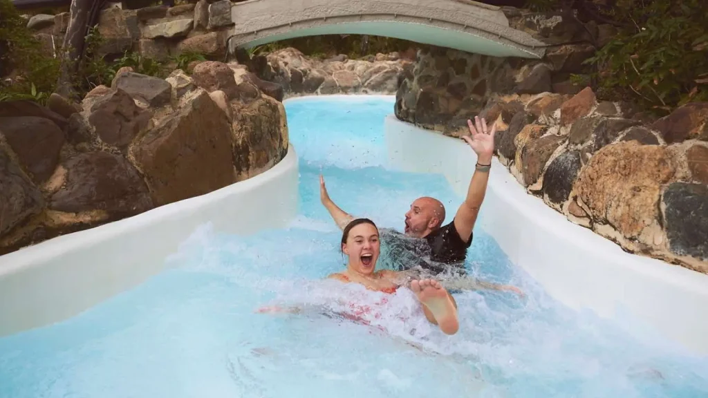 Two people riding down the Wild Water Rapids at the Subtropical Swimming Paradise at Center Parcs in Longleat water park, surrounded by rock features and splashing water.
