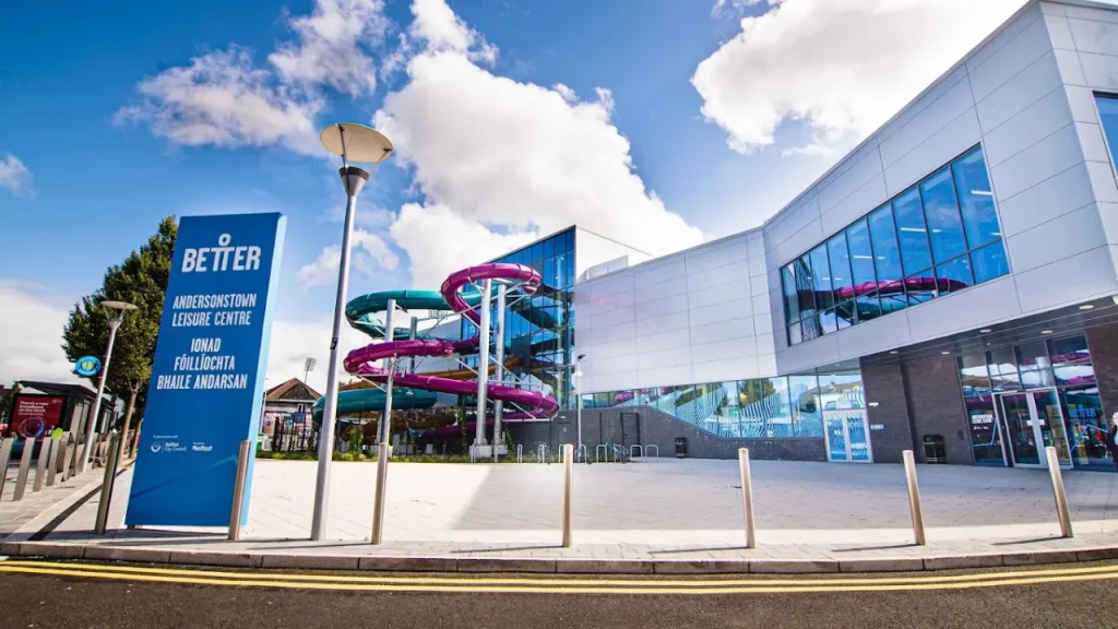 Exterior view of Andersonstown Leisure Centre in Belfast showing vibrant pink and teal spiral water slides and the modern entrance facade.