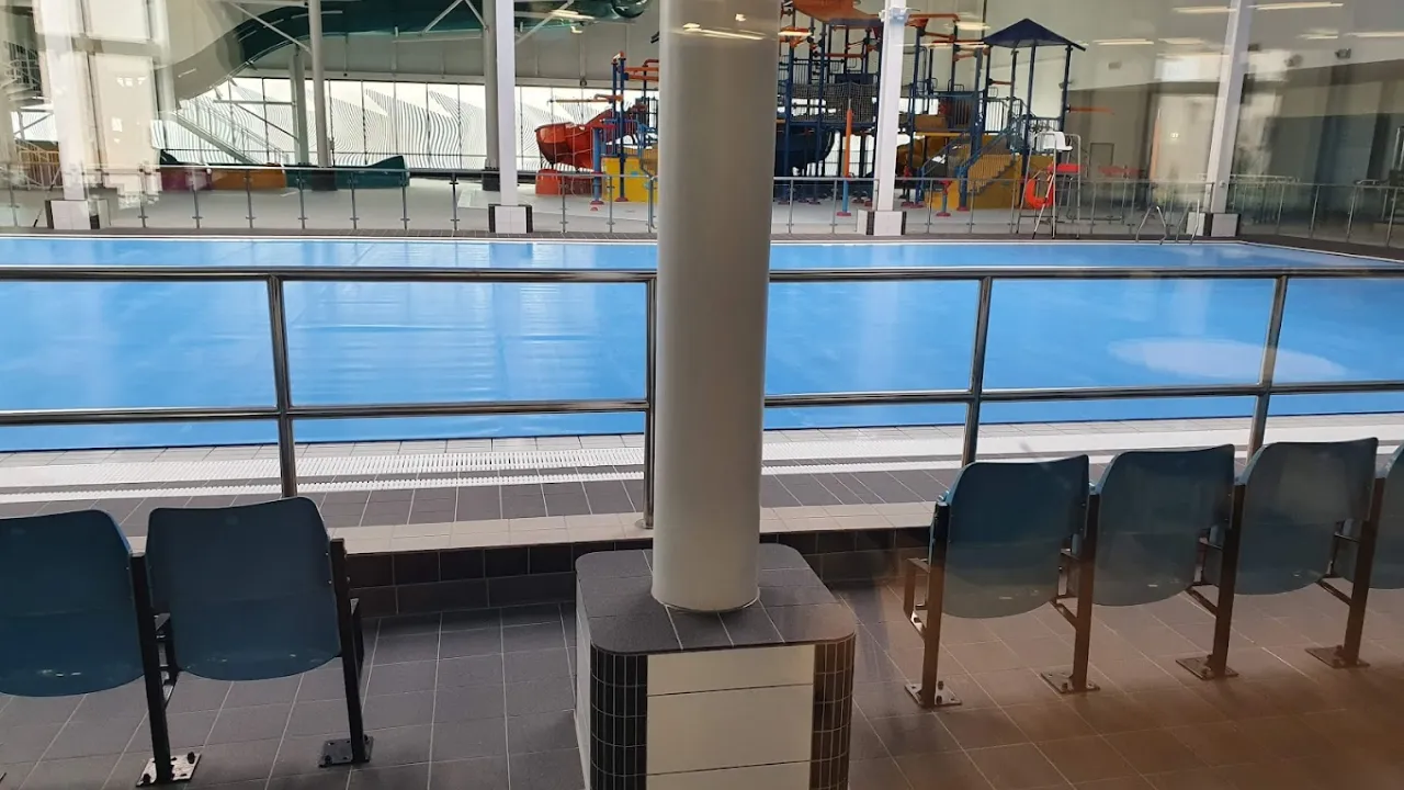 View of Andersonstown Leisure Centre’s pool from the spectator seating area, showing the calm water and children’s splash play structure in the background.