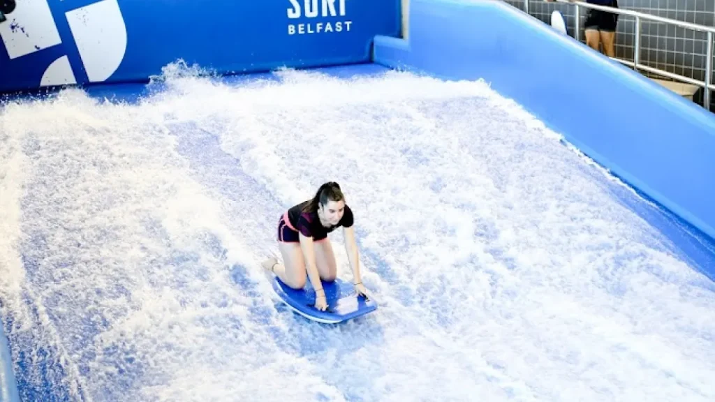 Girl bodyboarding on the Surf Belfast indoor surf simulator at Andersonstown Leisure Centre, surrounded by flowing water and safety barriers.
